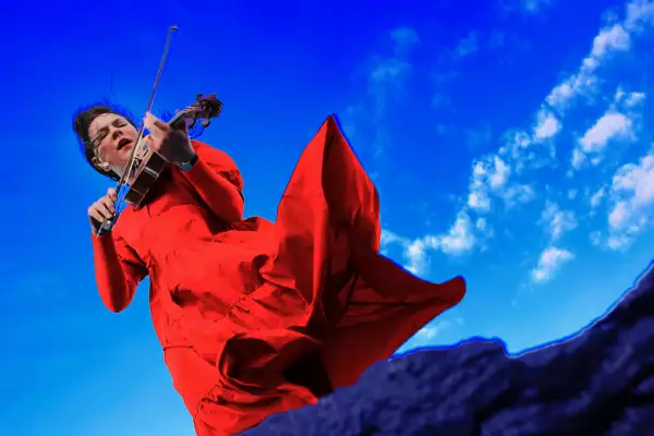 Clare Sands wearing a bright red dress and playing the fiddle, against a bright blue sky