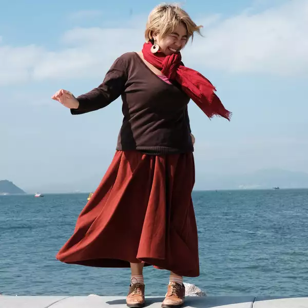 A woman stands in front of a body of water. Her arms are out by her sides as if in motion and her face is smiling, looking away from the camera. She wears a brown jumper, red scarf and orange-red skirt.