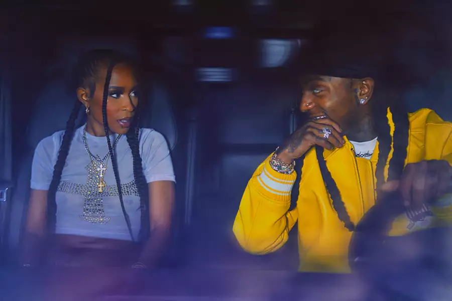Jacquees and Dej Loaf sit in the front seats of a car, facing each other. Dej Loaf wears a light blue crop top with a bejewelled cross, while Jacquees wears a yellow hoodie and smiles warmly.
