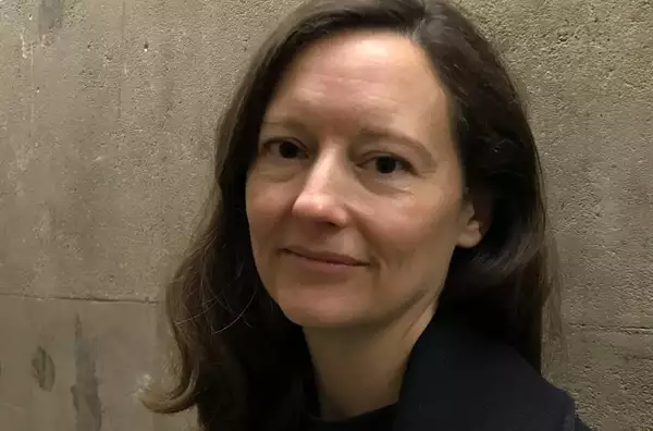 A close-up of a person standing against a grey wall. They have mid-length dark brown hair and dark eyes.