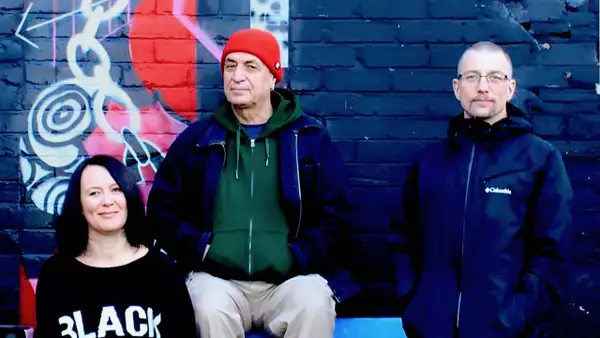 Three members of Oojami sit in front of a mural-painted brick wall with neutral expressions