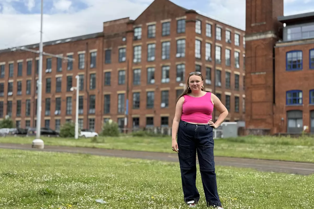 Ellie Brown wearing a bright pink top and jeans, stood outside a converted mill on the grass