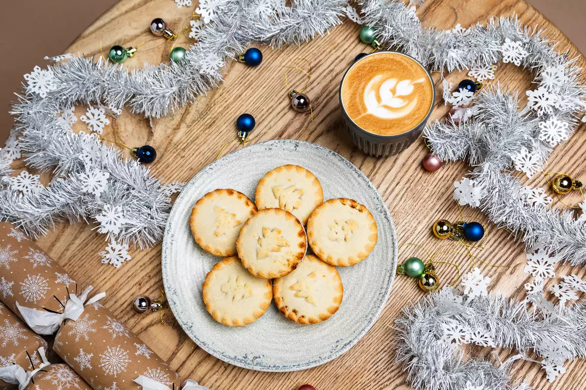 A festive table with six mince pies on a plate, a latte with leaf art, silver tinsel, snowflake cutouts, colorful baubles, and gift crackers.