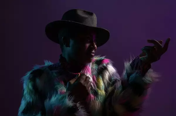 Jasmin Issaka - one of the Factory Sounds cohort for 2023 - wears a multicoloured faux fur jacket and black felt fedora a they pose against a purple background