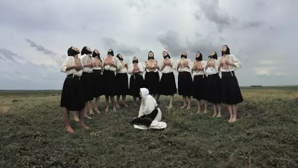 A group of women in traditional Eastern European attire stand barefoot in a grassy field under a dramatic, cloudy sky. Most are wearing black skirts, white blouses, and black headscarves, with some partially exposing their chests while lifting their heads upward in a ritualistic pose. In the foreground, a single woman dressed in white kneels on the ground with a headscarf and embroidered belt, gazing to the side.