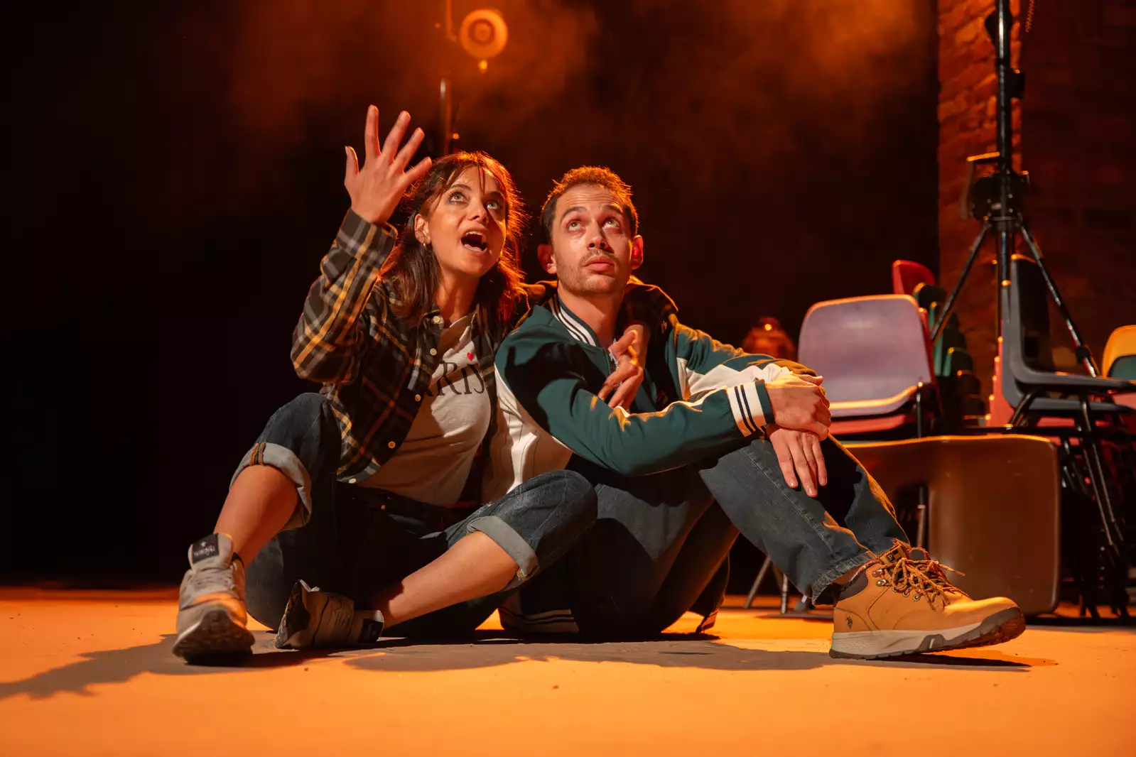 Two people sit on the floor of a stage staring up. One explains something to the other.