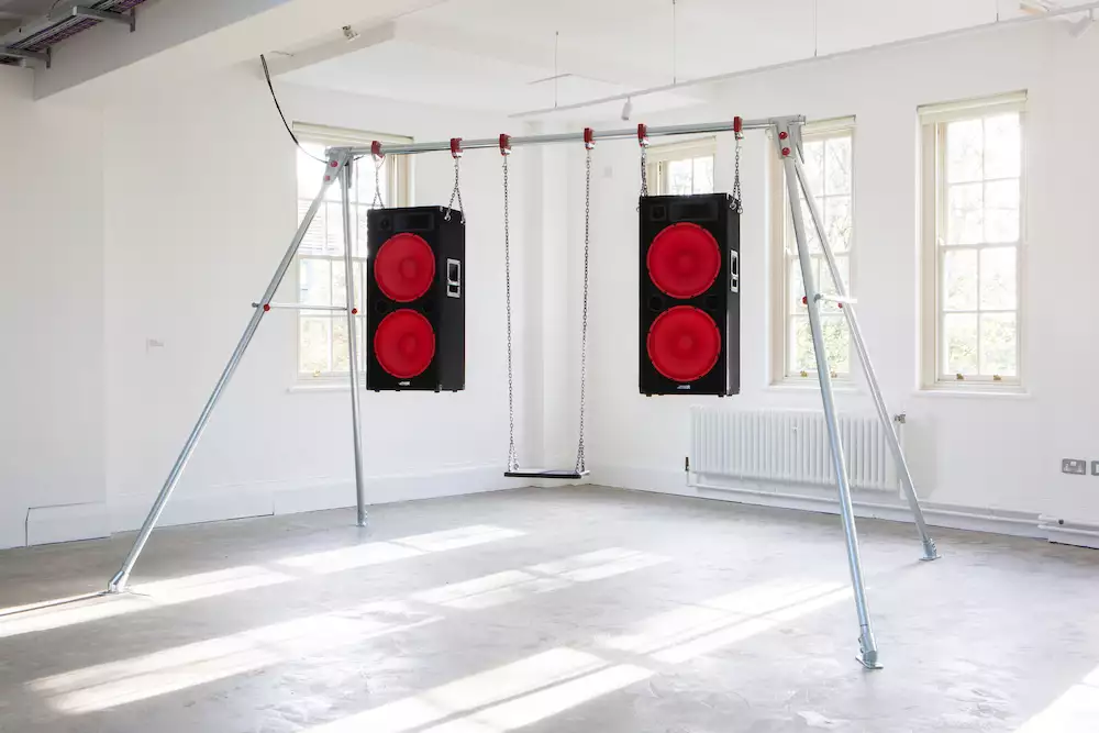 An artwork by Trackie McLeod showing a swingset with speakers hanging from it in a white gallery space