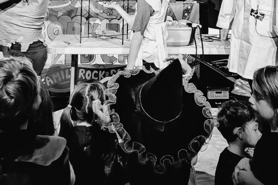 A black and white photo of children watching a performance. One is wearing a witch fancy dress hat.
