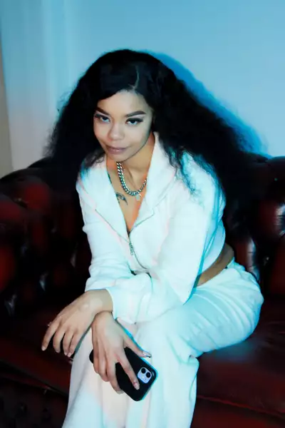 Tox6ix Scorpio sat on a sofa, holding a phone and wearing all white clothing