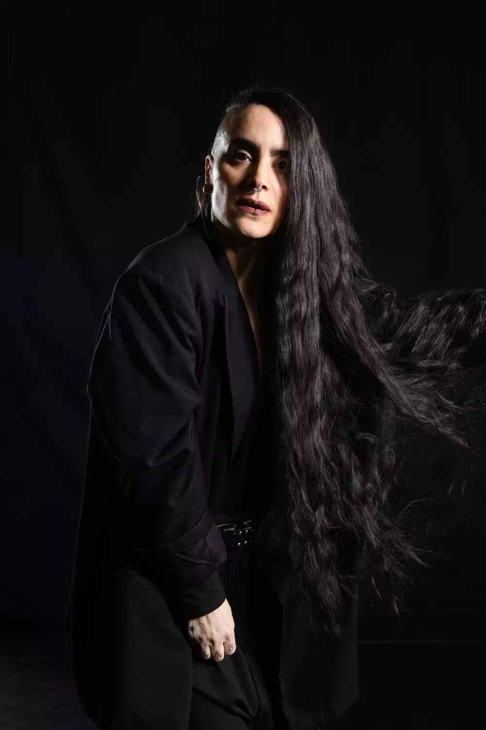 Portrait photo of Sonya Tayeh wearing all black, on a black background, flicking her long hair to the side