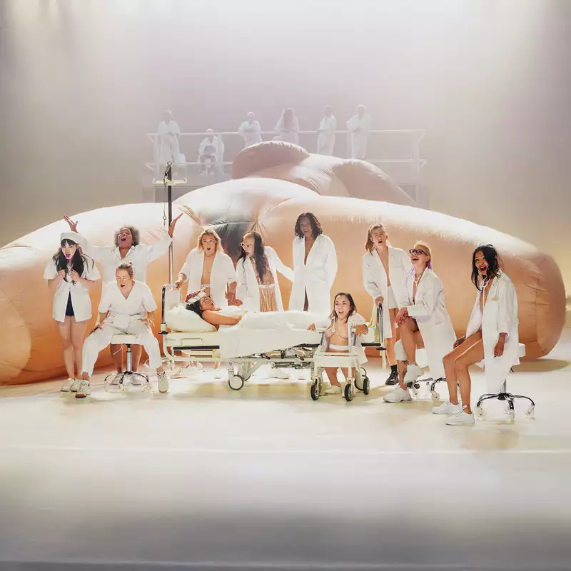 A group of performers on stage wearing white medical dress in front of a giant sculpture of a woman's body