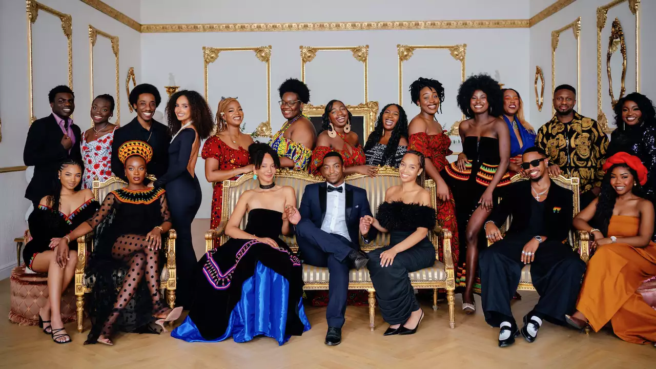 A large group of people pose together in a luxurious room with gold-trimmed walls and ornate furniture. They wear a mix of modern formalwear and vibrant traditional African attire, smiling warmly at the camera.