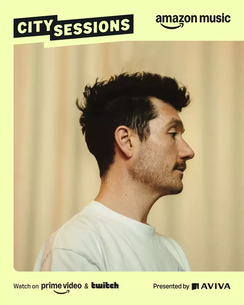 Dan Smith from Bastille in front of a softly lit beige curtain background. There is a yellow border with Amazon Music City Sessions Presented by Aviva in black text