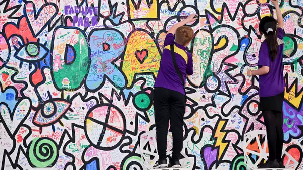 Two children in purple uniforms draw and write on a colorrful doodle wall filled with graffiti-style art and messages.