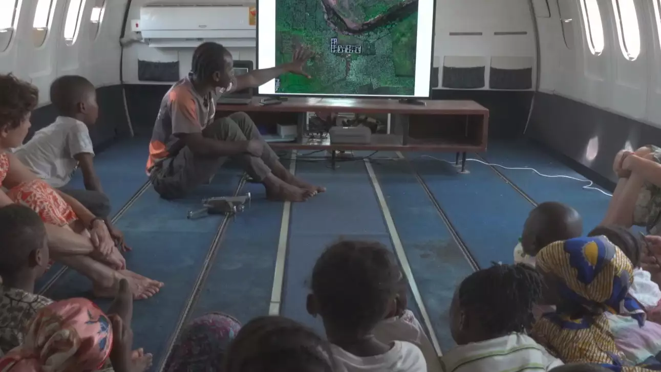 A photograph of a man gesturing at a TV screen showing a map, while delivering a lesson to a group of children in Ghana. The lesson is taking place in a retired airplane with the rows of seats removed.