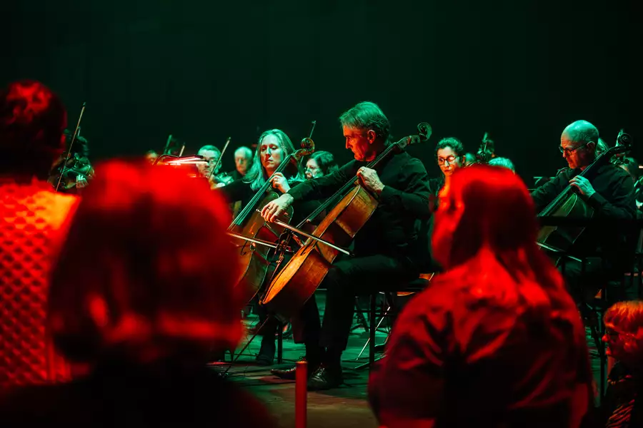 Two audience members watching a cello player in an orchestra. The lighting is red.