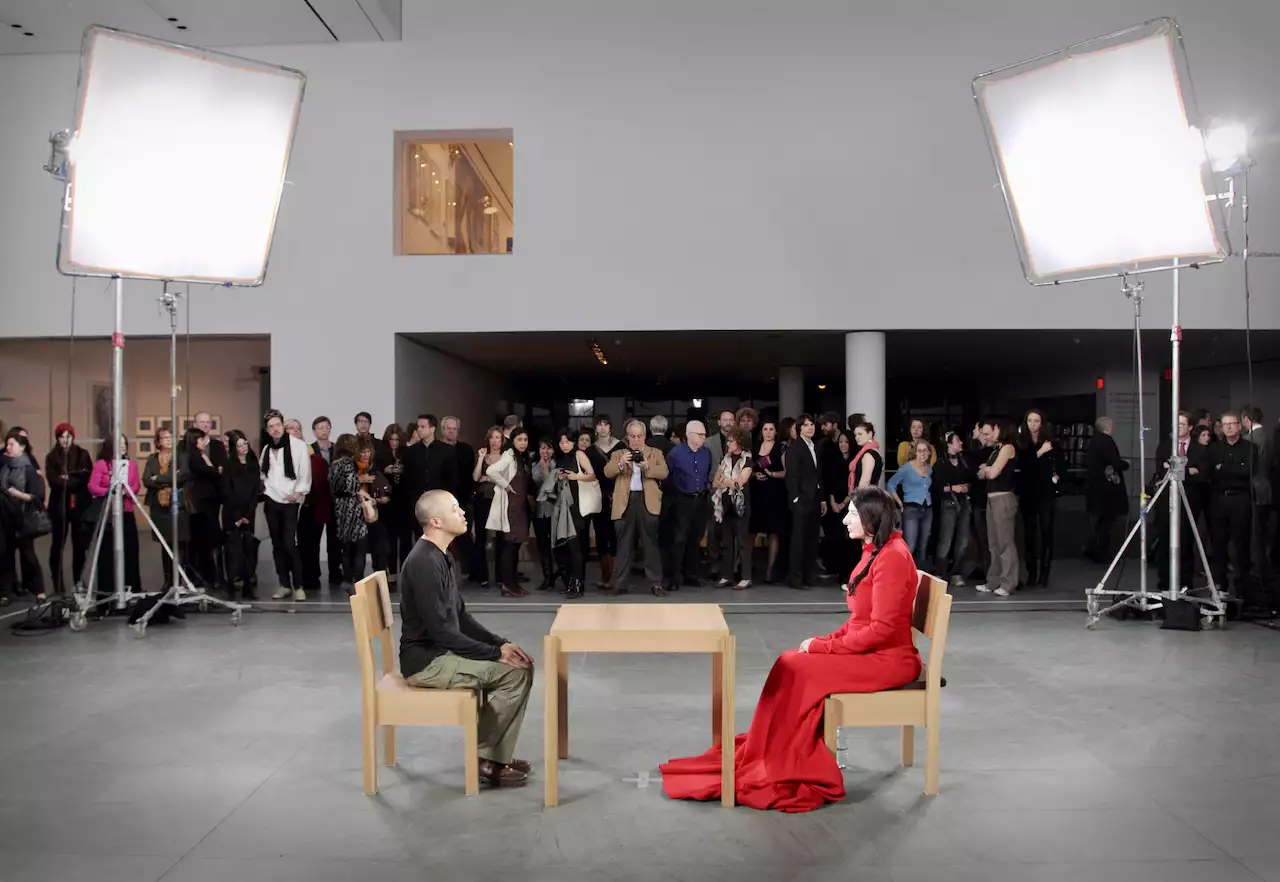 Marina Abramović seated across from an audience member during The Artist is Present at MOMA. She is wearing a red dress. There is a crowd watching.