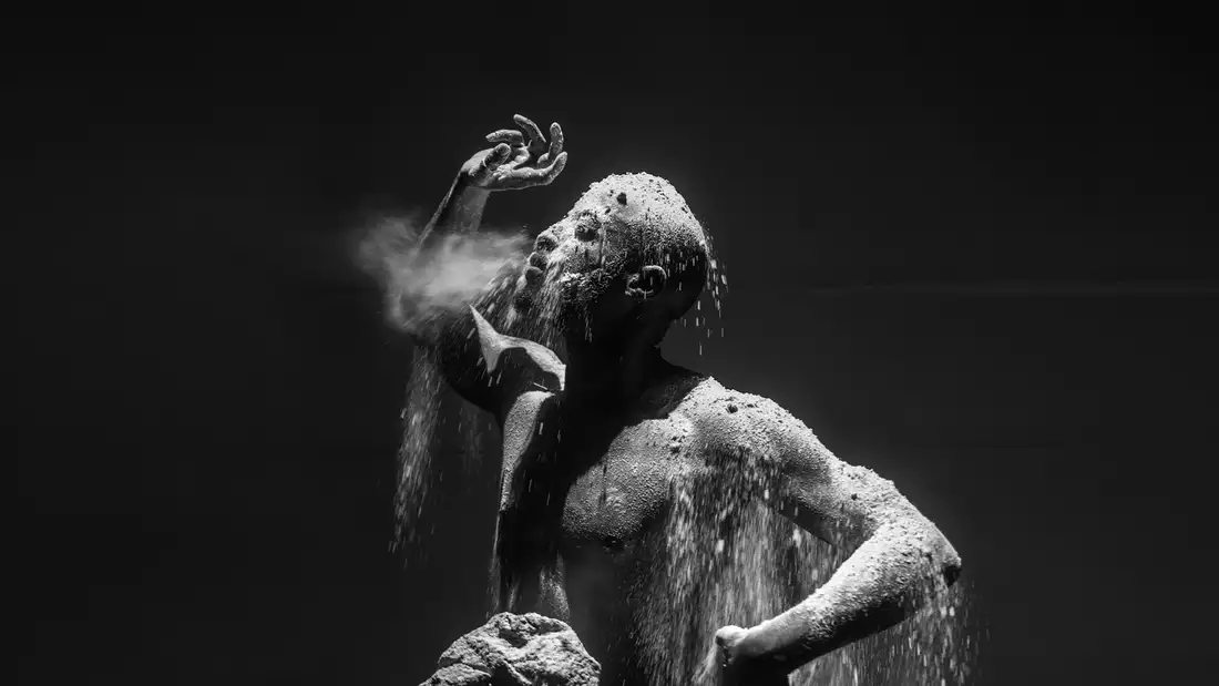 A dramatic black-and-white photograph of a man covered in dust and sand, with particles cascading over his body as he exhales forcefully.