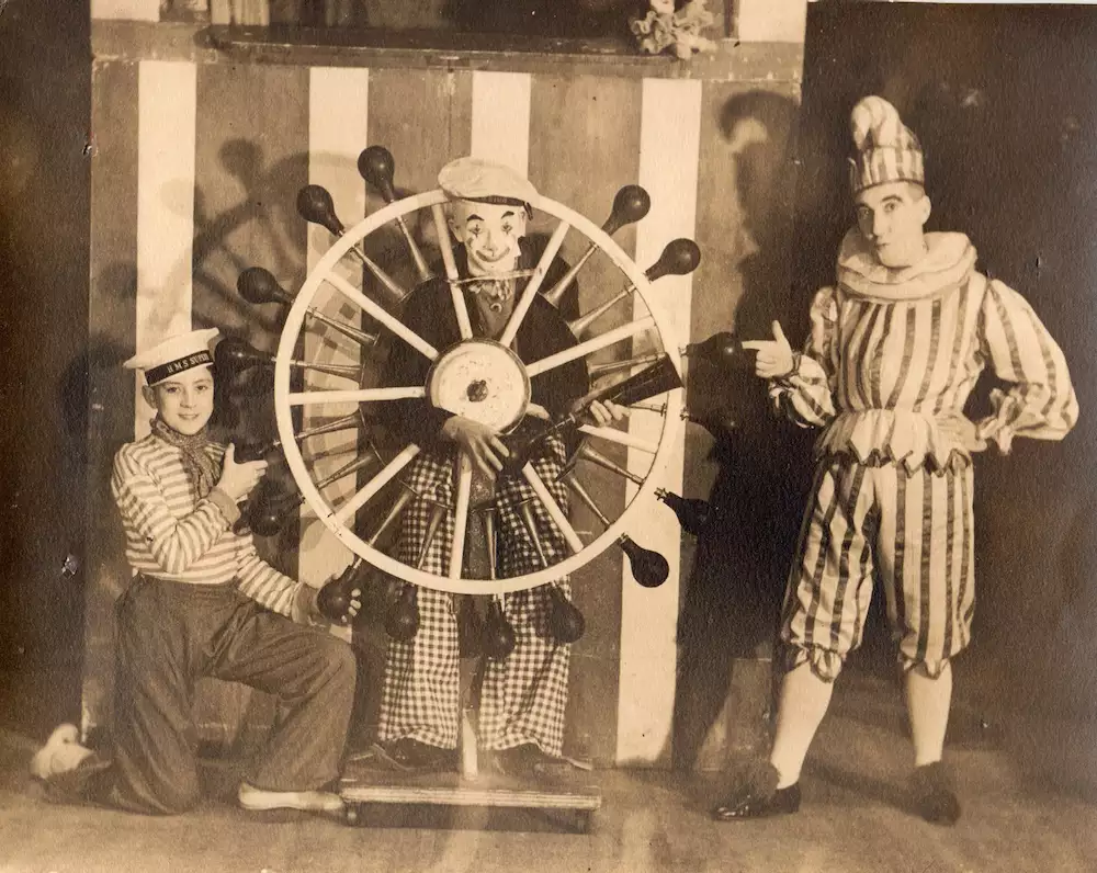 Vintage photo of three people in theatrical costumes, one framed inside a large ship’s wheel while the others point at him.