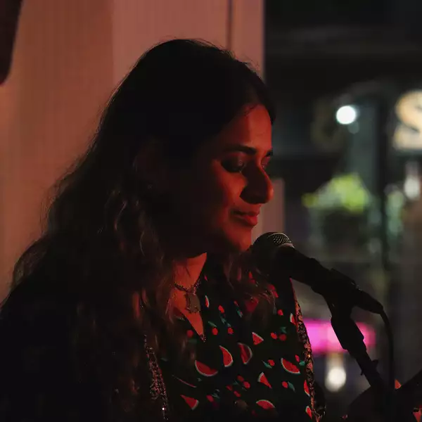 afshan d’souza-lodhi reading into a microphone wearing a top with watermelons on it. The background is dark.