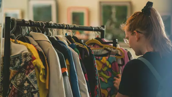 A person looking through a rail of brightly coloured clothes. Their hair is tied back with a black scrunchie and they have a blue rucksack on.