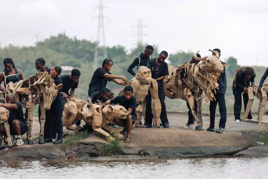 A herd of cardboard animal puppets being operated by people in black clothes by the side of a river