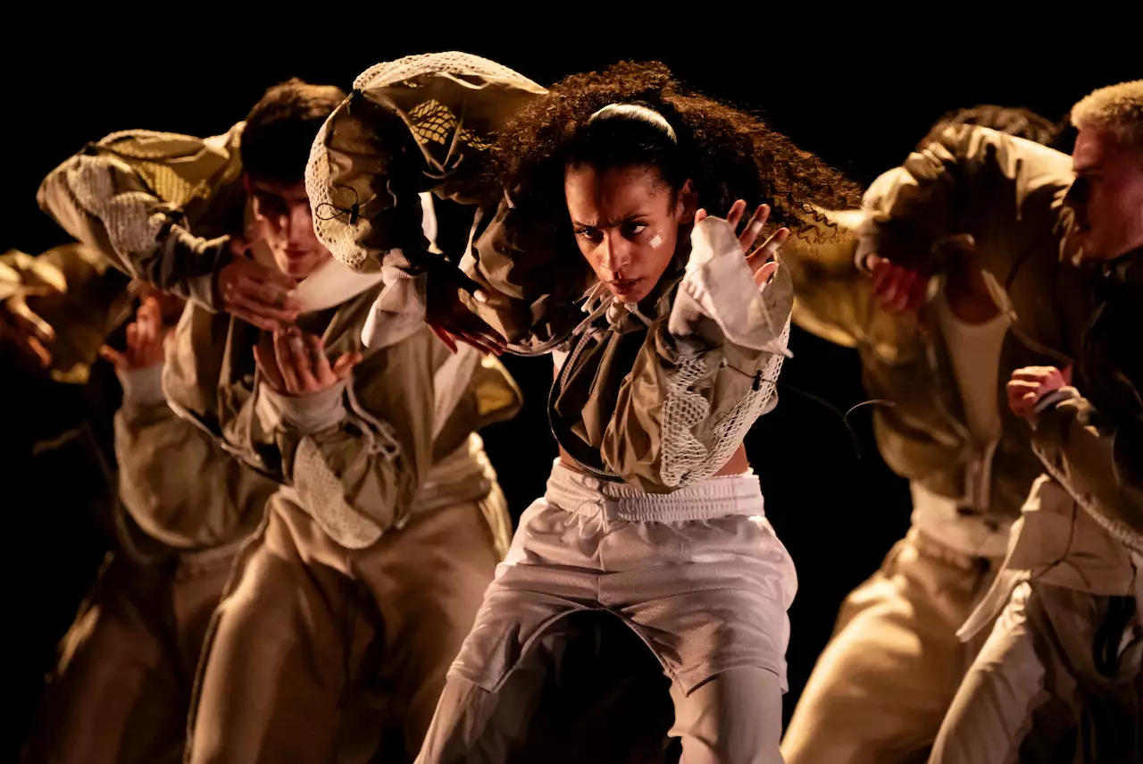 A group of dancers in coordinated white outfits performing under warm, dramatic lighting as part of Cycles.