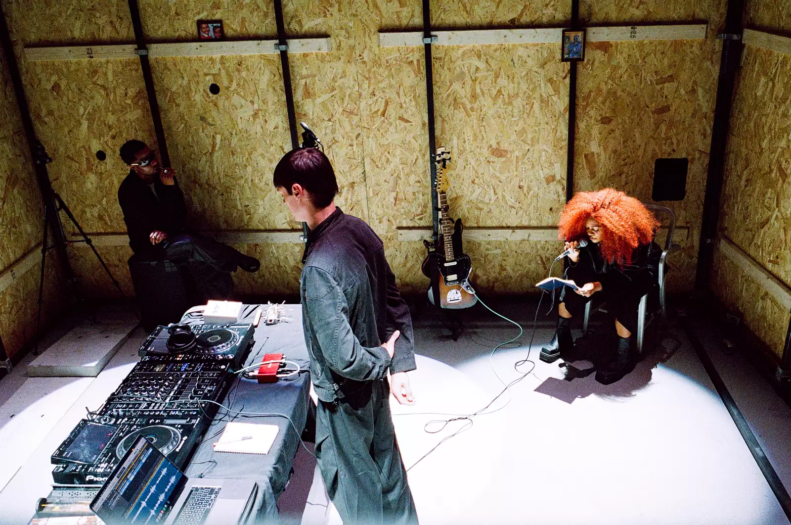Three members of 54: Manchester performing inside a large crate with a guitar, microphone and decks