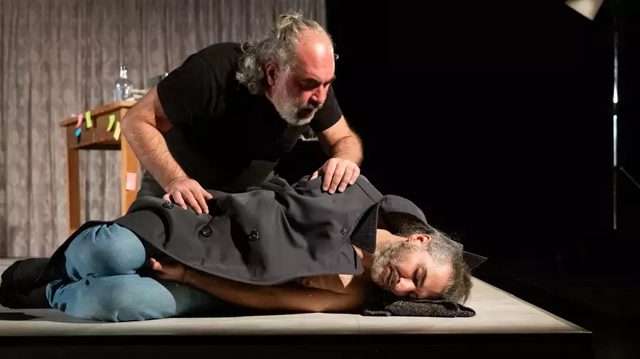 A person lying down on stage with eyes closed underneath a coat – with another person peering over him