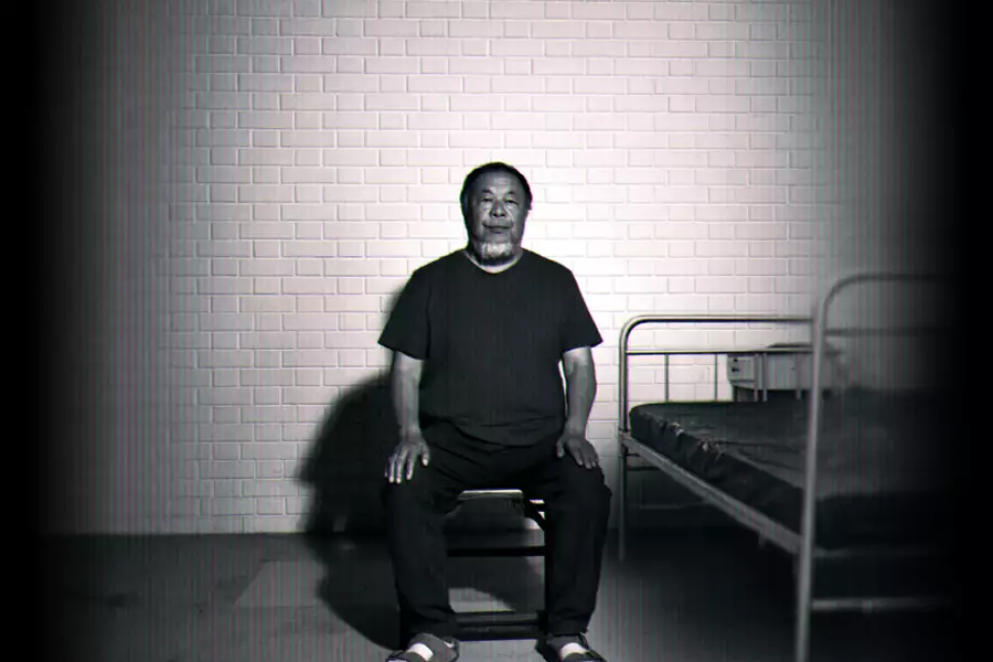 Black-and-white image of Ai Weiwei sitting on a chair beside a simple metal bed against a brick wall.