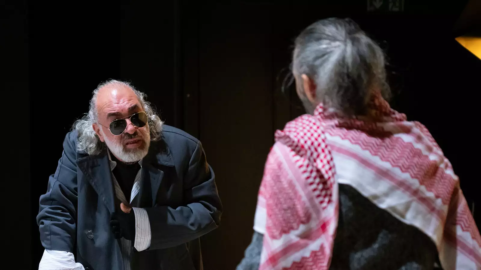 On stage, a person in sunglasses and a big coat speaks to another person with his back turned