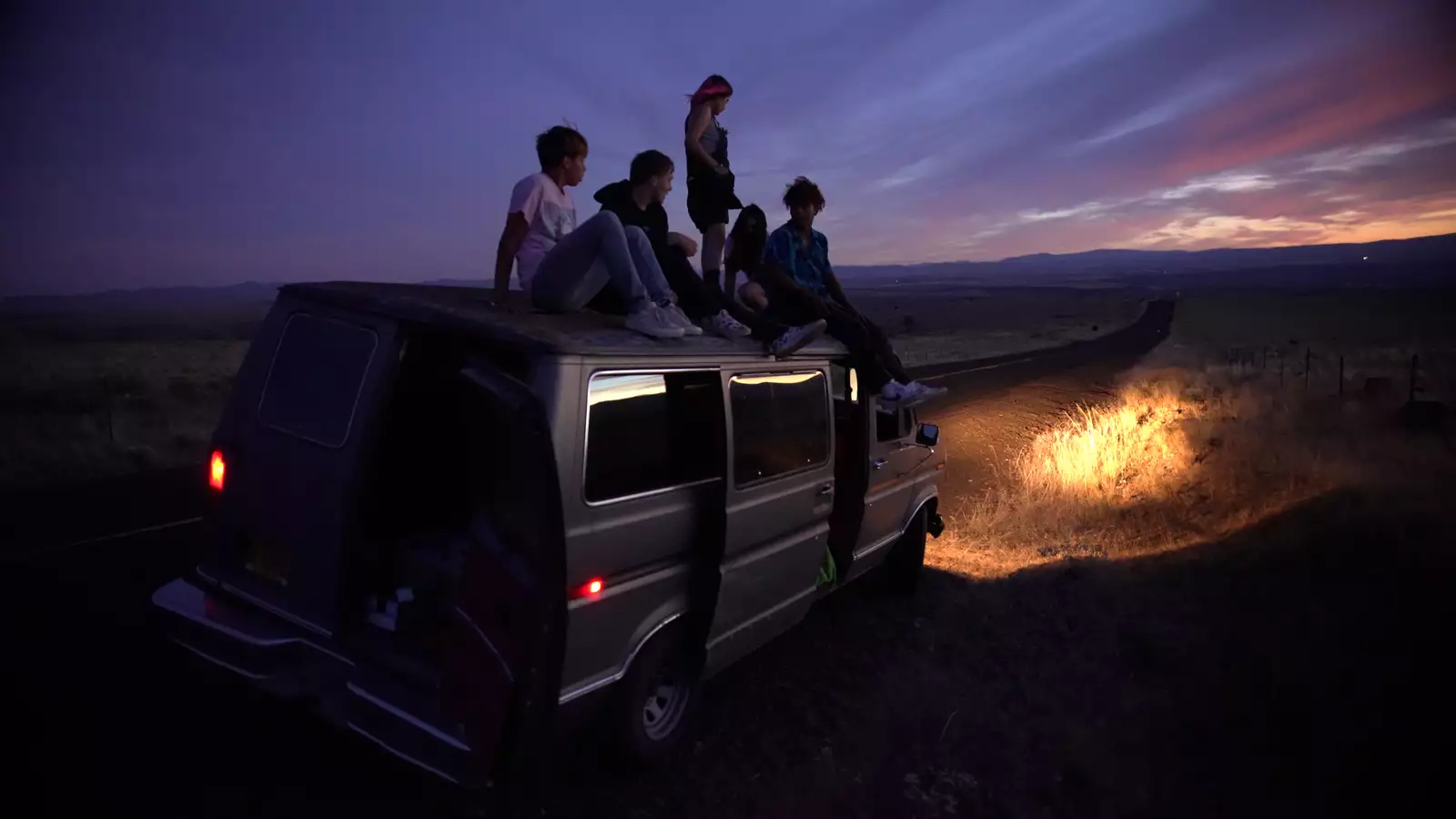 Still from Gasoline Rainbow. Five young people sat on top of a van off the side of a road. The sun is setting.