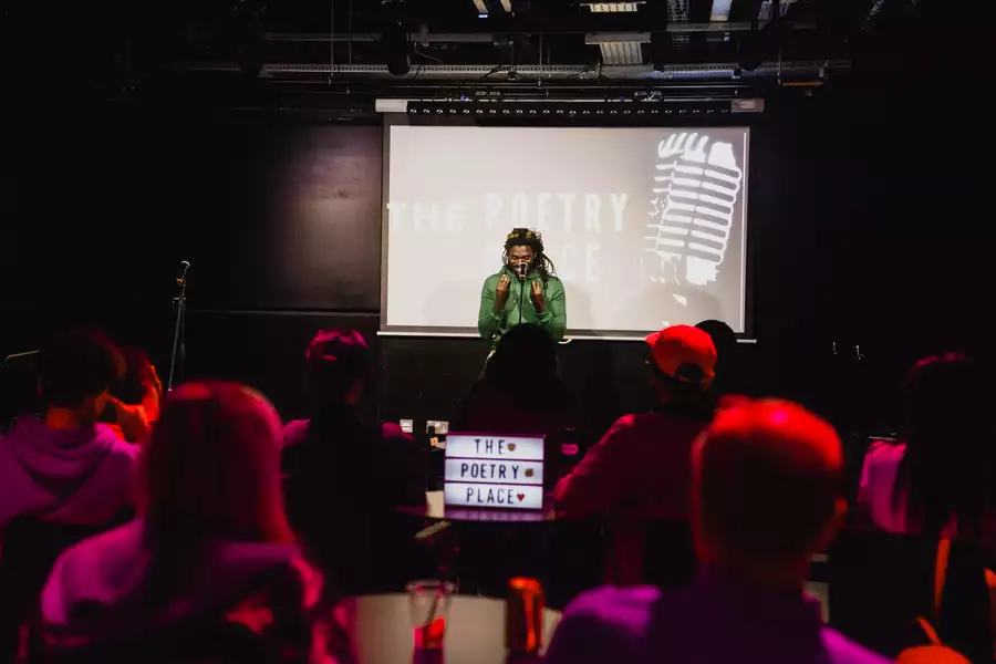 A poet performs on stage at a spoken word event as an audience watches in a dimly lit venue. There is a screen behind them which says 'poetry place'.