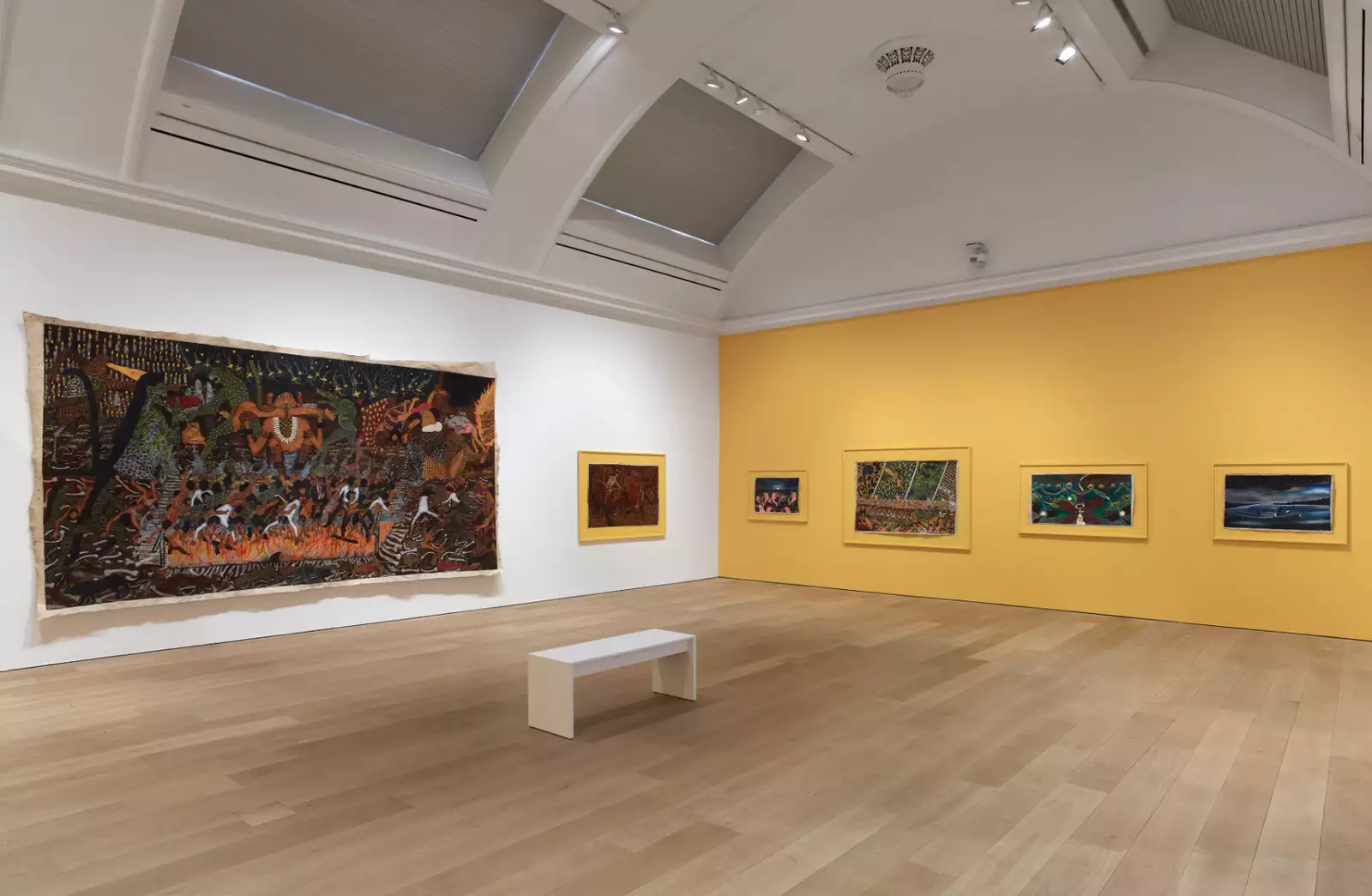 A photo of a gallery with a white wall and a yellow wall with 4 small paintings and one large by Santiago Yahuarcani