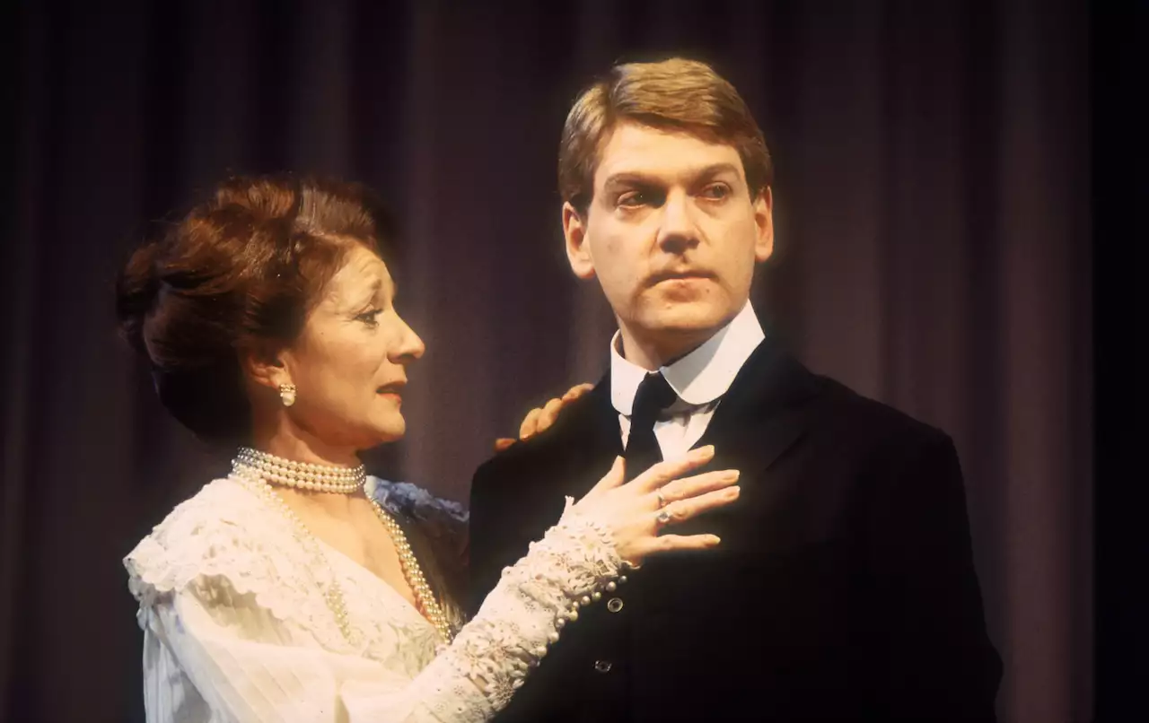 Jane Lapotaire and Kenneth Branagh on stage as Gertrude and Hamlet. Jane is wearing a white lace dress with her hands on Kenneth's chest. He is wearing a suit and looking off-camera.