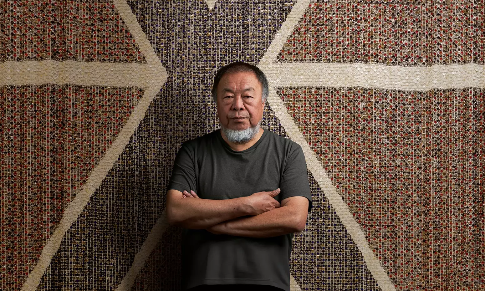 Ai Weiwei stood with arms folded wearing a black t-shirt, in front of a giant flag made of buttons
