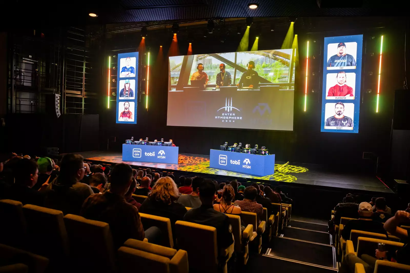 Enter Atmosphere esports event with players on stage in the Hall, audience watching, and large screens displaying game and commentators.