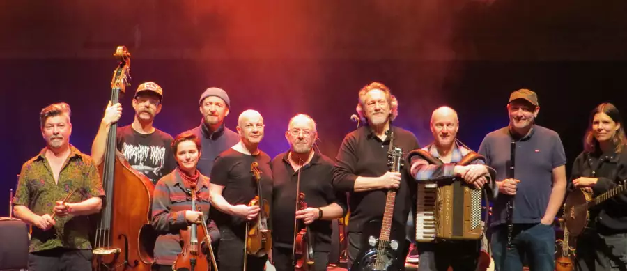 A group of musicians stand together on stage holding their instruments, including fiddles, guitar, accordion, flute, upright bass, and banjo. Warm red stage lighting creates a lively atmosphere behind them.