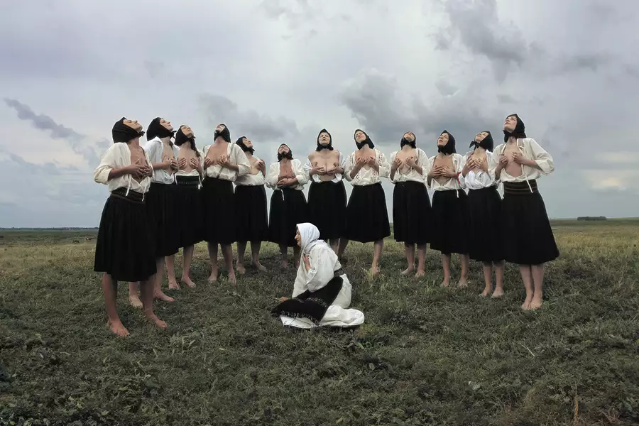 A group of women in traditional Eastern European attire stand barefoot in a grassy field under a dramatic, cloudy sky. Most are wearing black skirts, white blouses, and black headscarves, with some partially exposing their chests while lifting their heads upward in a ritualistic pose. In the foreground, a single woman dressed in white kneels on the ground with a headscarf and embroidered belt, gazing to the side.