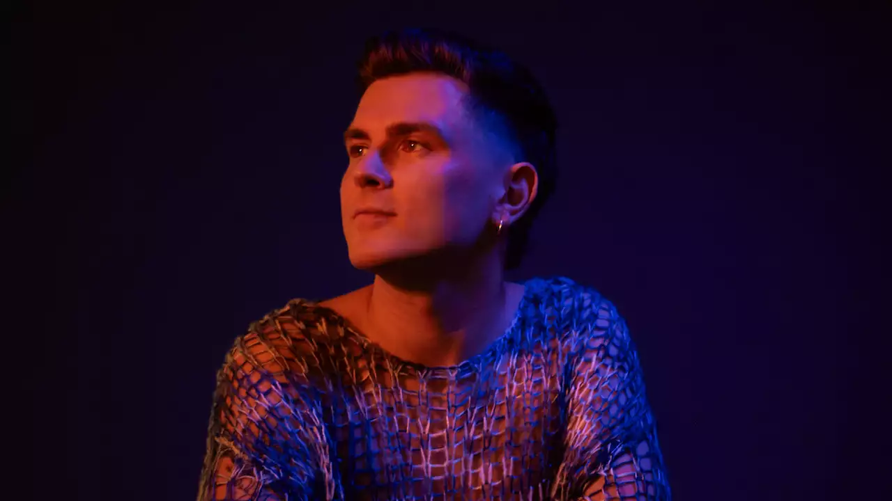 Fastlove wearing a meshed colourful top, looking to the side. The lighting is red.