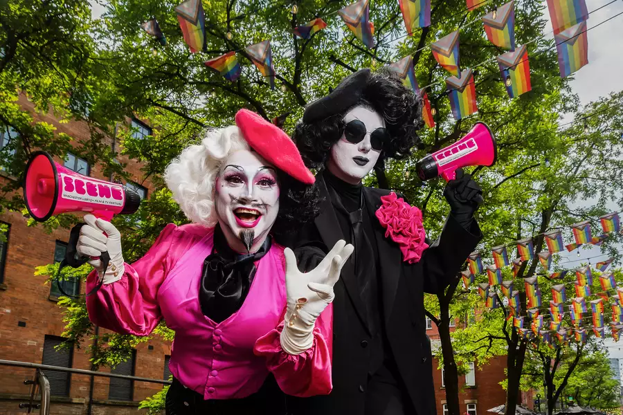 Anna Phylactic and Liquorice Black pose on Manchester's Canal Street. There are colourful pride flags above them. They are holding pink branded microphones.