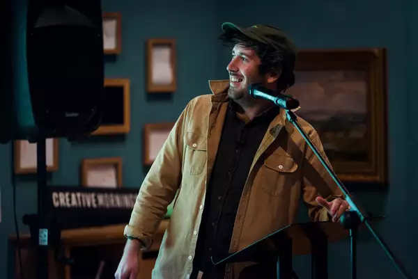 A man wearing a beige shirt and green cap stands in front of a microphone. In the background, picture frames are visible on a teal-coloured wall.