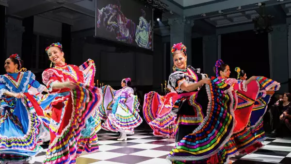 A group of women in colorful traditional Mexican dresses perform a folkloric dance on a black-and-white checkered floor, smiling and swirling their vibrant skirts.