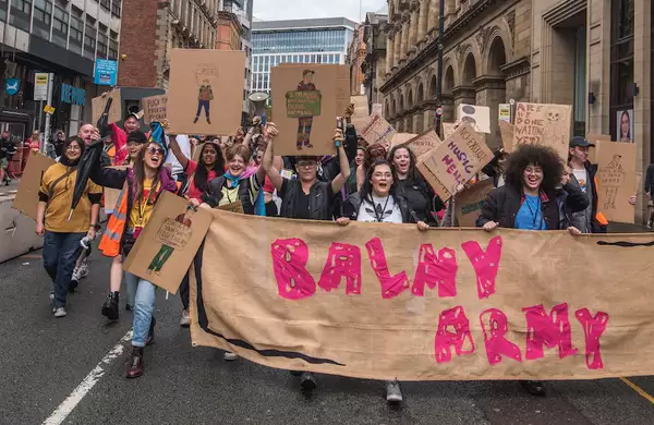 A group of young people marching through Manchester City Centre with a huge banner that reads 'Balmy Army'. Many are holding placards and megaphones.