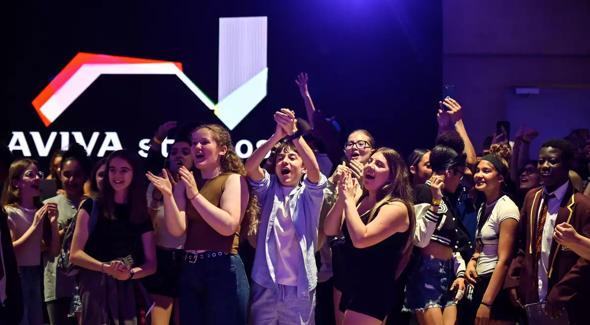 A cheering crowd of young people celebrate enthusiastically in the Social, with a glowing "Aviva Studios" sign in the background.