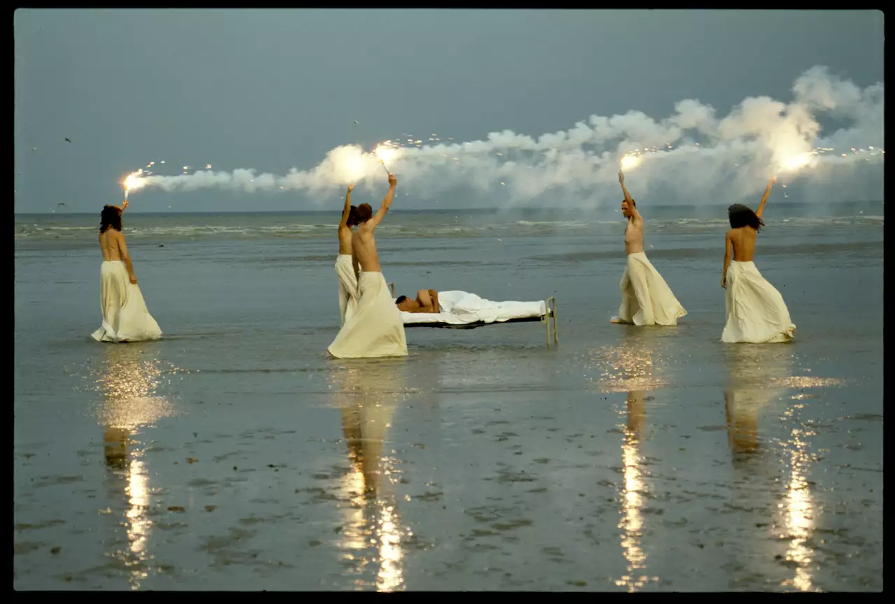Still from Derek Jarman's The Garden (1990), showing five people stood in a circle, wearing white and holding flares on the sea shore. In the middle a man lies in a bed.