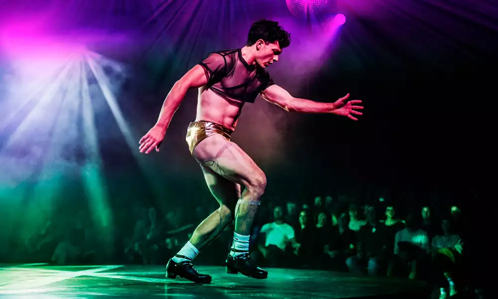 A dancer performs on stage during WAKE, wearing gold shorts and a sheer black crop top, illuminated by colorful stage lights.