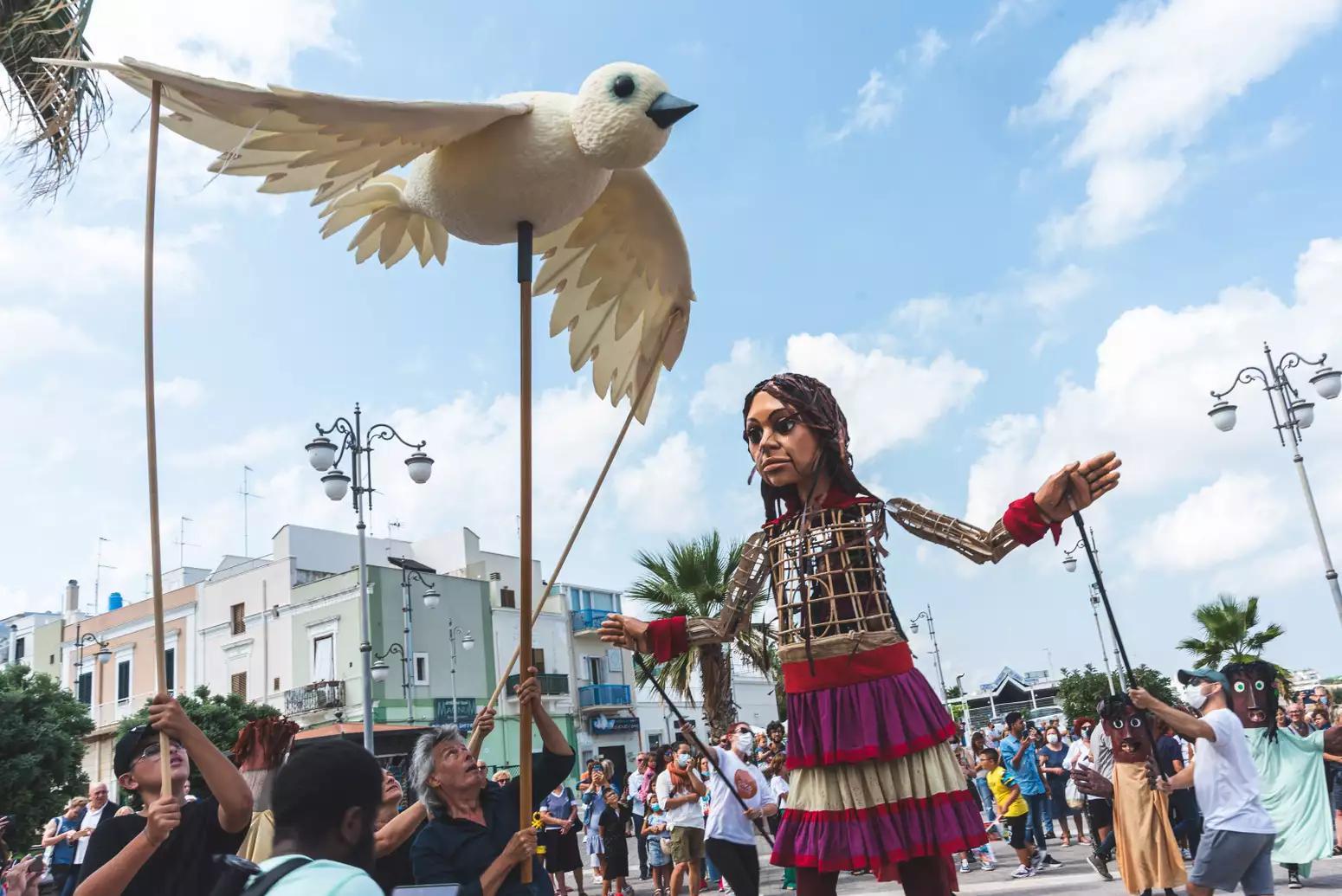 The Little Amal puppet stretches her arms out, mimicking the big white bird flying front of her that's held up by 3 performers holding sticks. Behind her is the city centre, a crowd and 2 performers with papier-mâché heads.