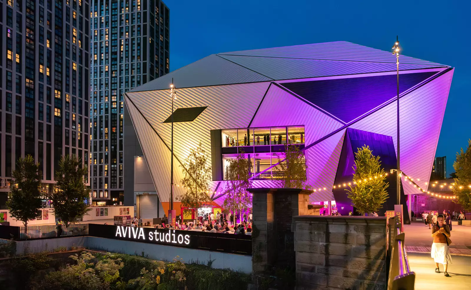 A photo of the venue Aviva Studios lit up with purple lights with crowds enjoying Festival Square by the river