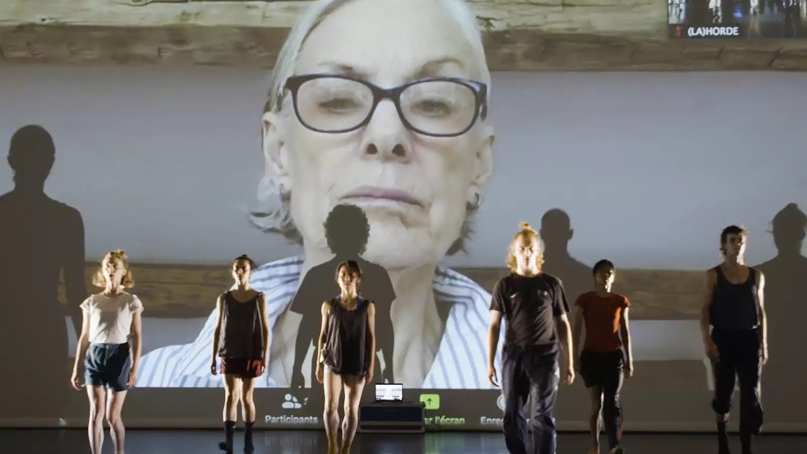 A photograph of 6 dancers from LA(HORDE), standing at attention in front of a background projection showing a Zoom call with Lucinda Childs